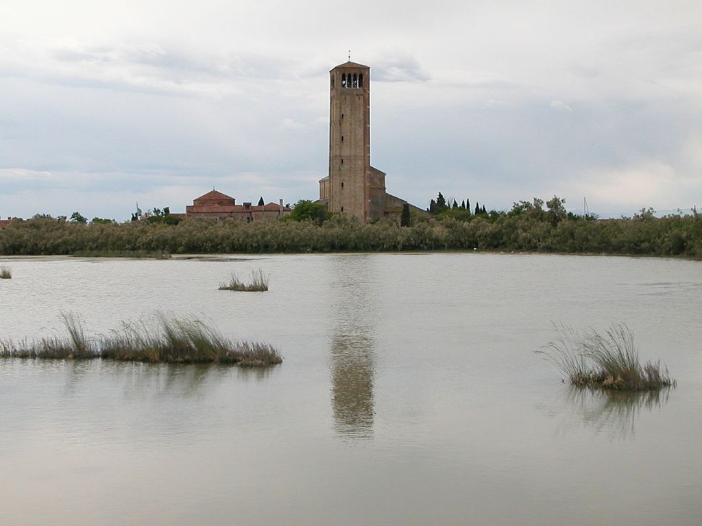 View of the bell tower and buildings of Torcello island reflected in the Venetian lagoon with marshland in the foreground.