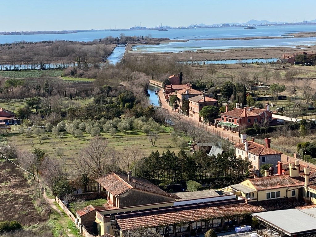 Aerial view of the island of Torcello in the Venetian Lagoon, showcasing green fields, residential buildings with red rooftops, and waterways leading to the lagoon in the background.