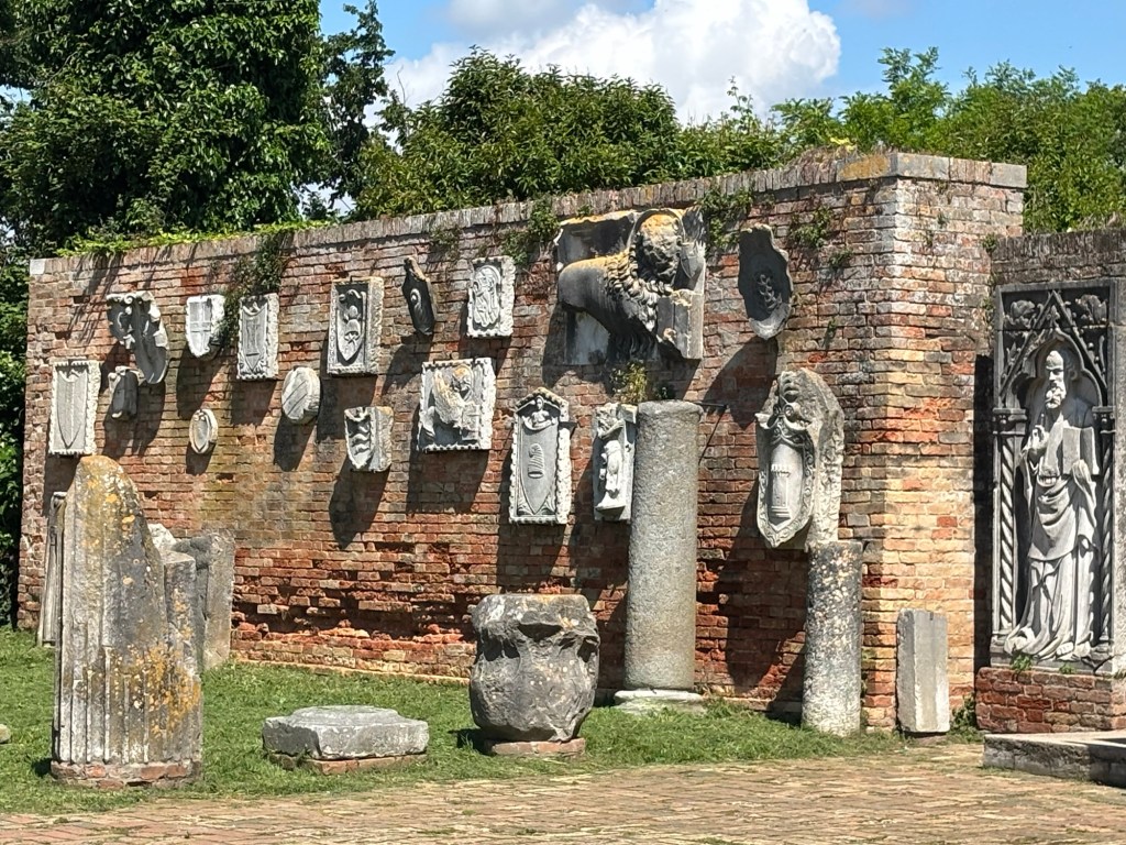 An outdoor display of ancient stone artifacts, including sculptures and bas-reliefs mounted on a brick wall, surrounded by greenery.