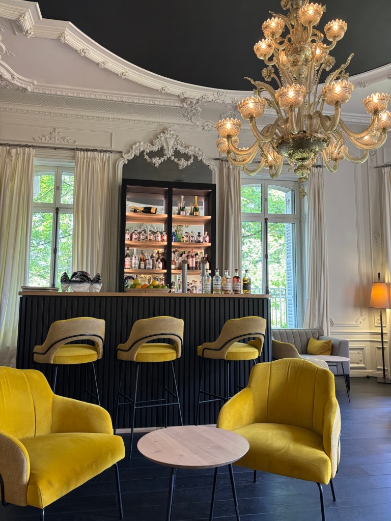 Interior of a stylish bar at Chateau de Beaulieu, featuring yellow velvet chairs, a wooden bar with various bottles on display, and elegant chandeliers against a beautifully detailed ceiling.