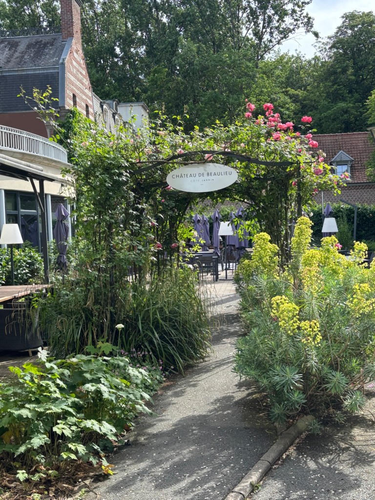 Entrance to Chateau de Beaulieu framed by lush greenery and blooming roses, with outdoor seating visible under shaded patio umbrellas.