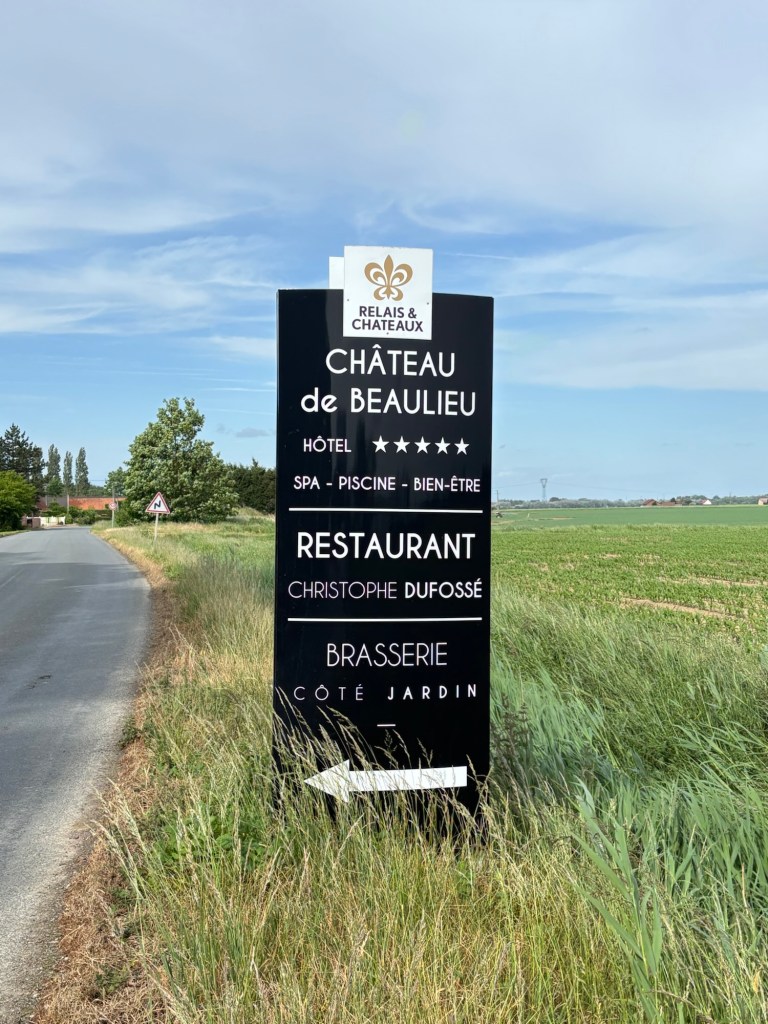 Sign indicating the direction to Chateau de Beaulieu, a five-star hotel and restaurant, surrounded by green fields under a clear sky.