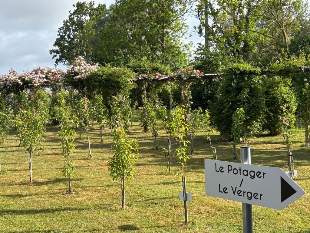 A well-maintained garden with trees and flowering plants, featuring a sign pointing to 'Le Potager / Le Verger' amidst lush greenery.